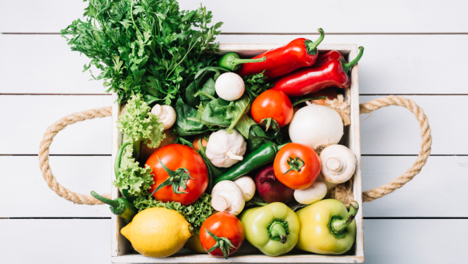 Dārzeņi (tomāti, paprika, salāti) kastē ar rokturiem, fonā balti dēļi