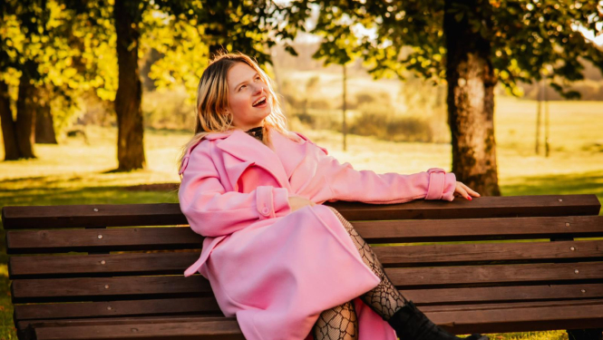 Meitene sēž uz soliņa rozā mētelī, skatās labajā stūrī, smaida