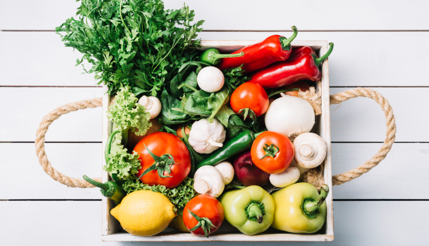 Dārzeņi (tomāti, paprika, salāti) kastē ar rokturiem, fonā balti dēļi